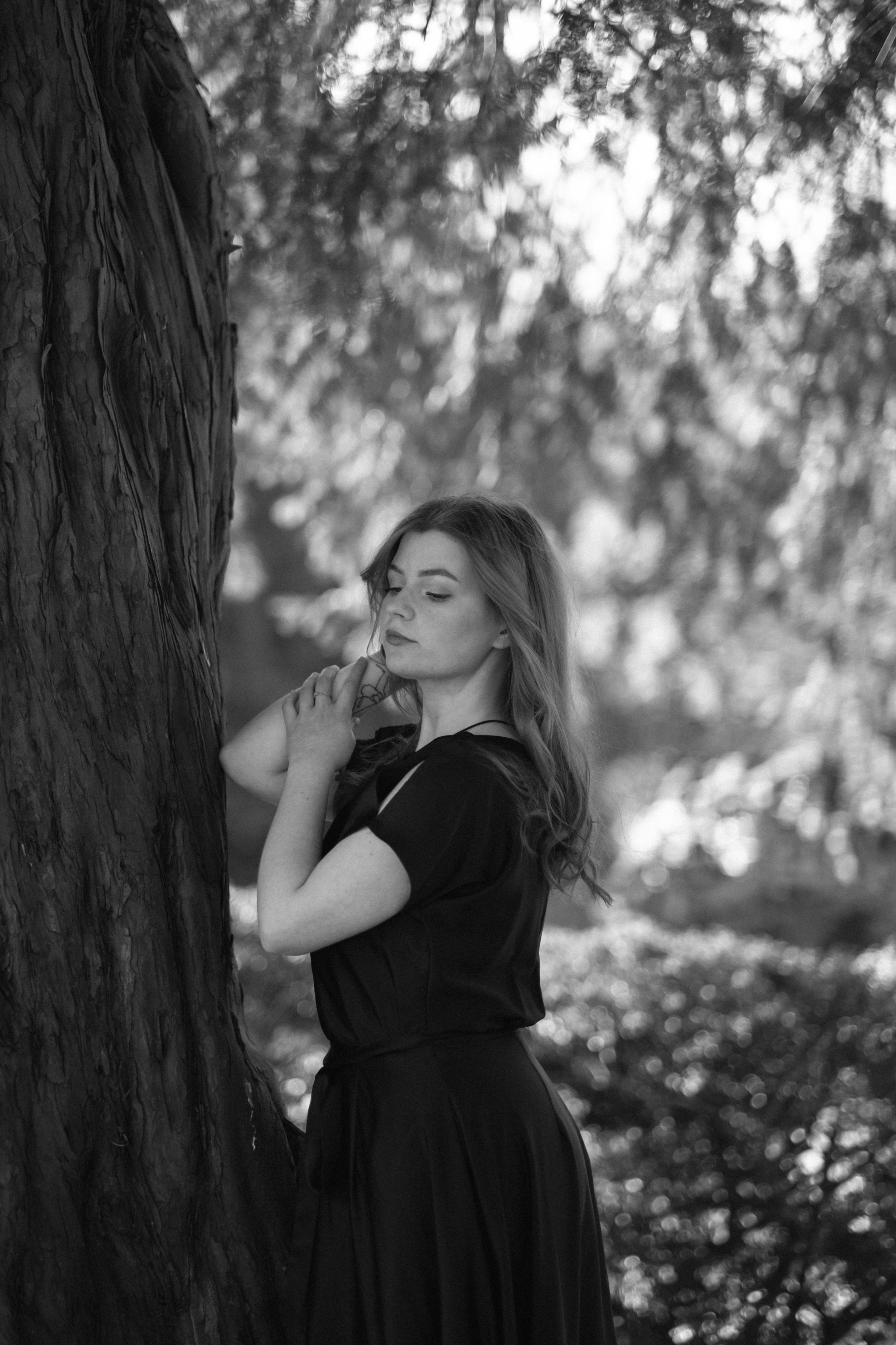 A woman in a black dress leans against a tree in a serene park setting.
