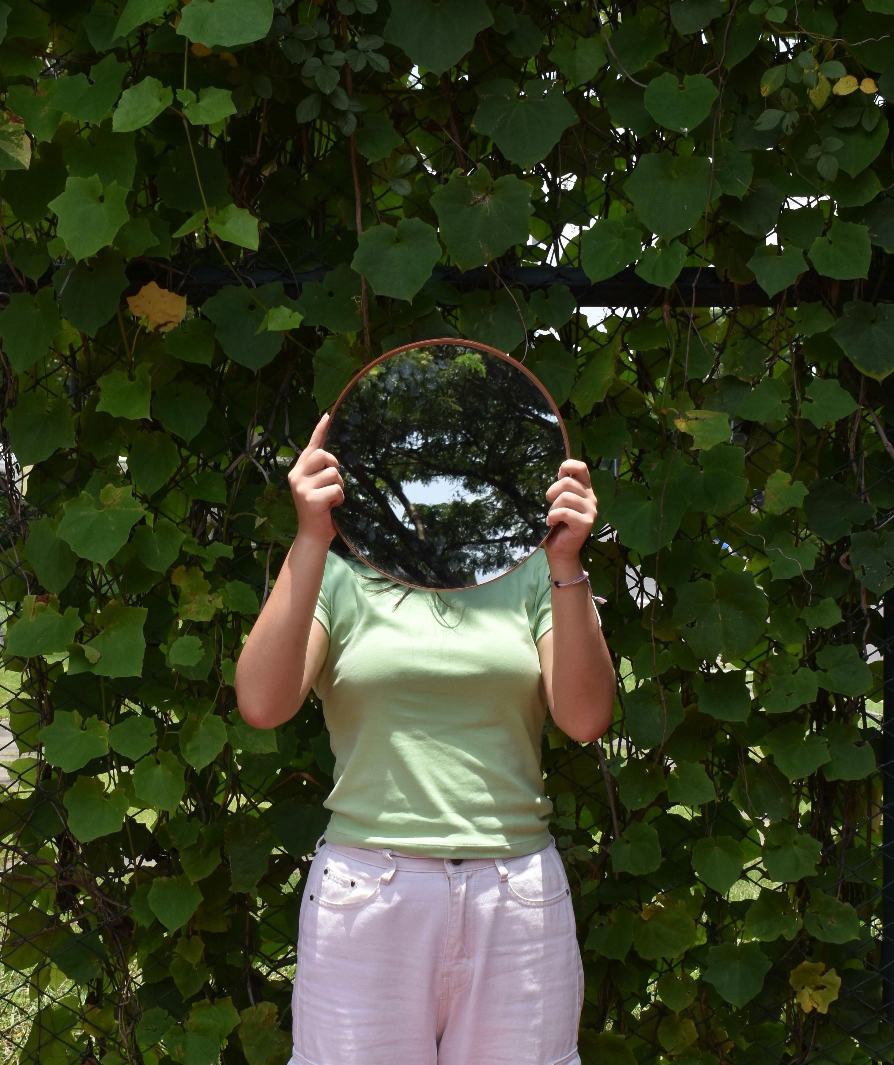 A woman stands in front of lush greenery holding a mirror reflecting trees and sky.