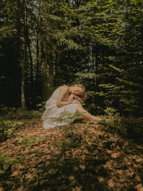 A woman in a white dress is sitting on the ground in the woods