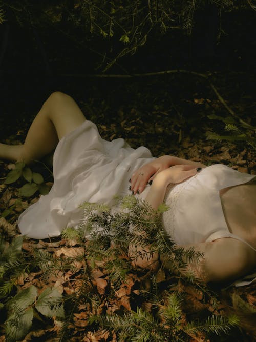 A woman in a white dress laying on the ground in the woods