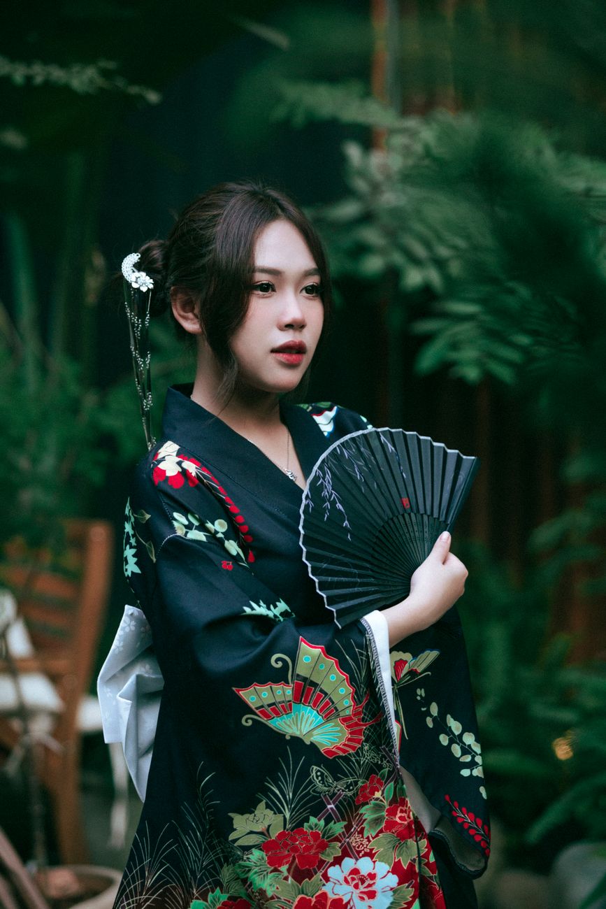 A portrait of a woman in a traditional Japanese kimono, elegantly holding a fan amidst greenery.
