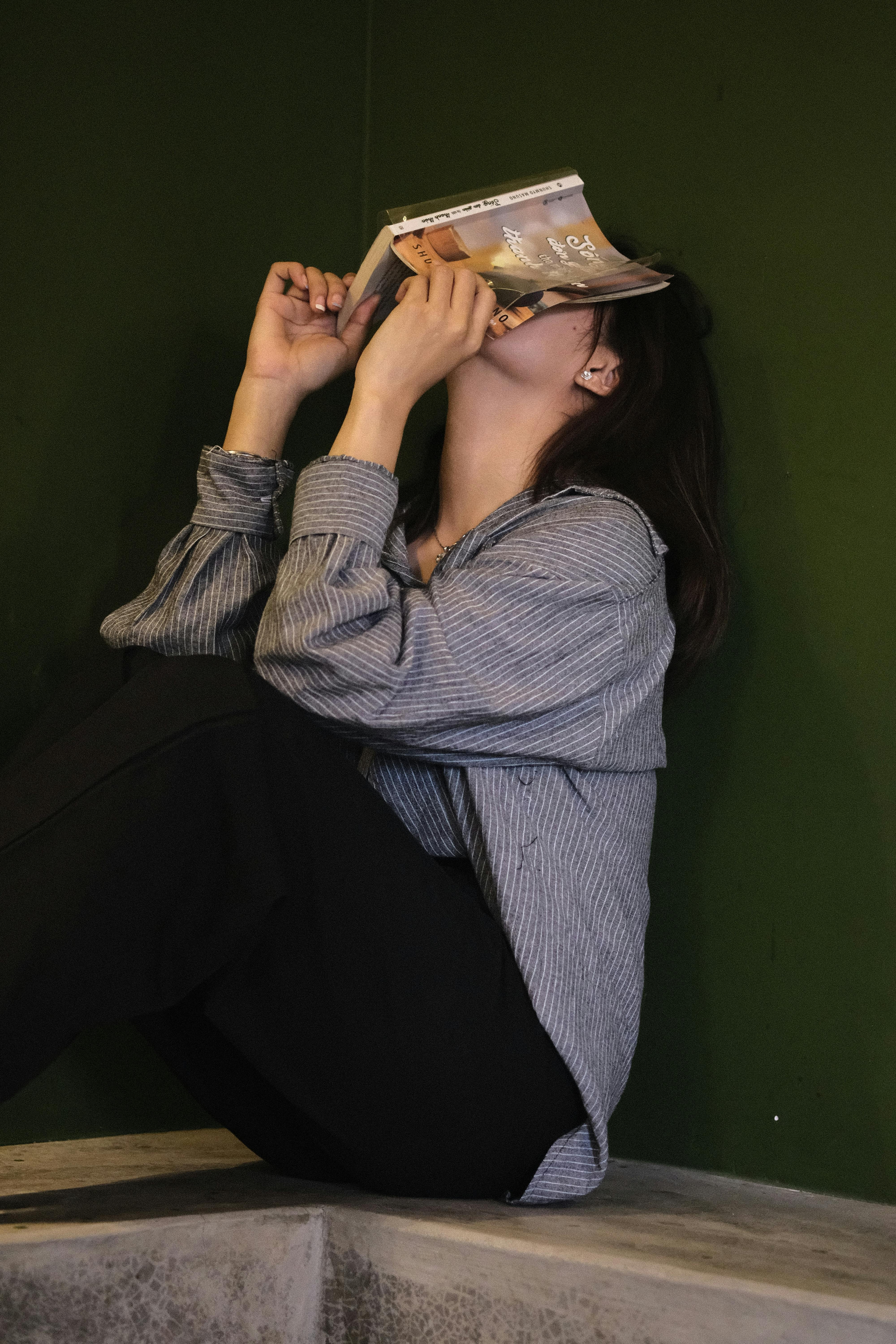 Free Woman sitting and relaxing with a book against a green wall, creating a cozy indoor vibe. Stock Photo