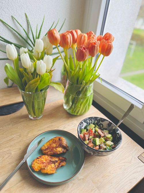Free A cozy dining setup with colorful tulips, grilled chicken, and salad on a wooden table by the window. Stock Photo