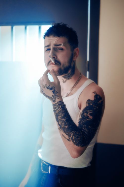 Free Portrait of a tattooed man in a white tank top, posing with a cigarette indoors Stock Photo