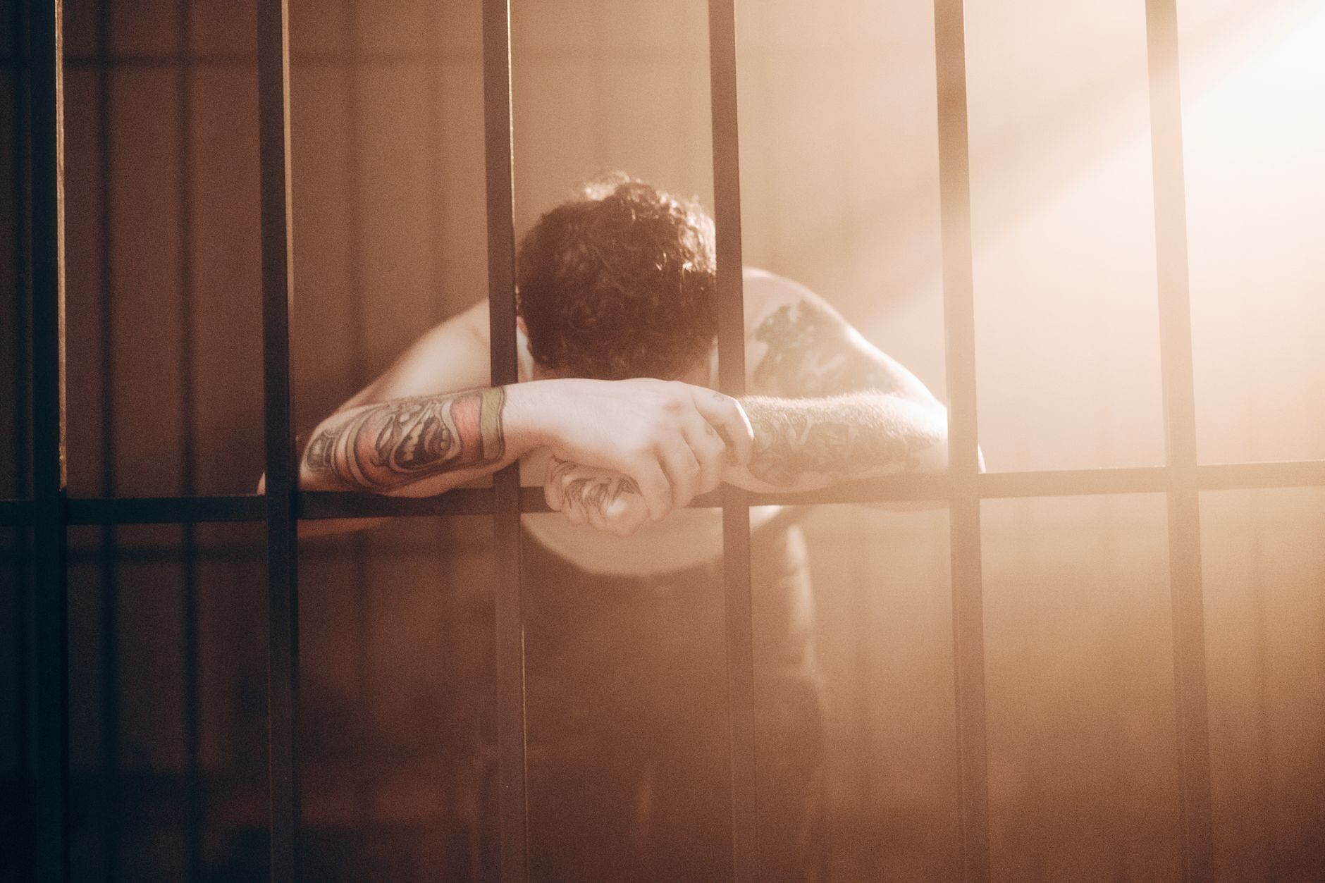 A tattooed man leans against prison bars, illuminated by dramatic sunbeams. Moody photography.
