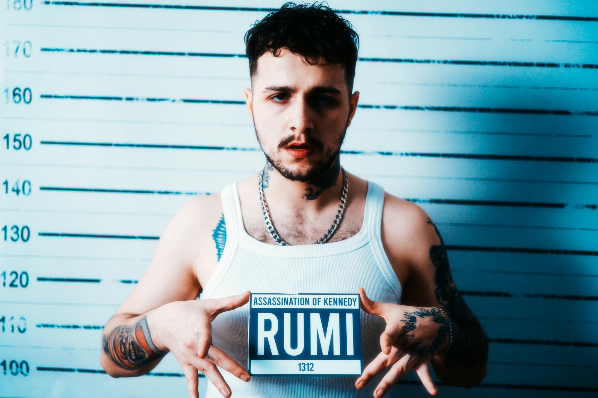 Portrait of a tattooed man holding a sign against a mugshot backdrop.