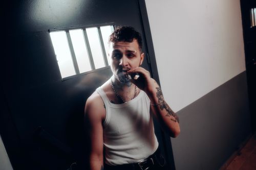 Free Adult man in tank top smokes cigarette near barred window, evoking prison theme. Stock Photo