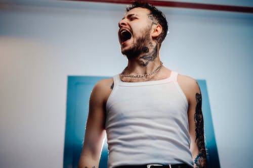 A man with tattoos and a white tank top is screaming