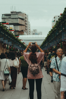 A woman takes a photo of a bustling city street filled with people and shops, capturing urban life.