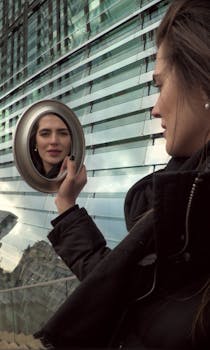A young woman in a black coat gazes at her reflection in a round mirror with a modern building in the background.