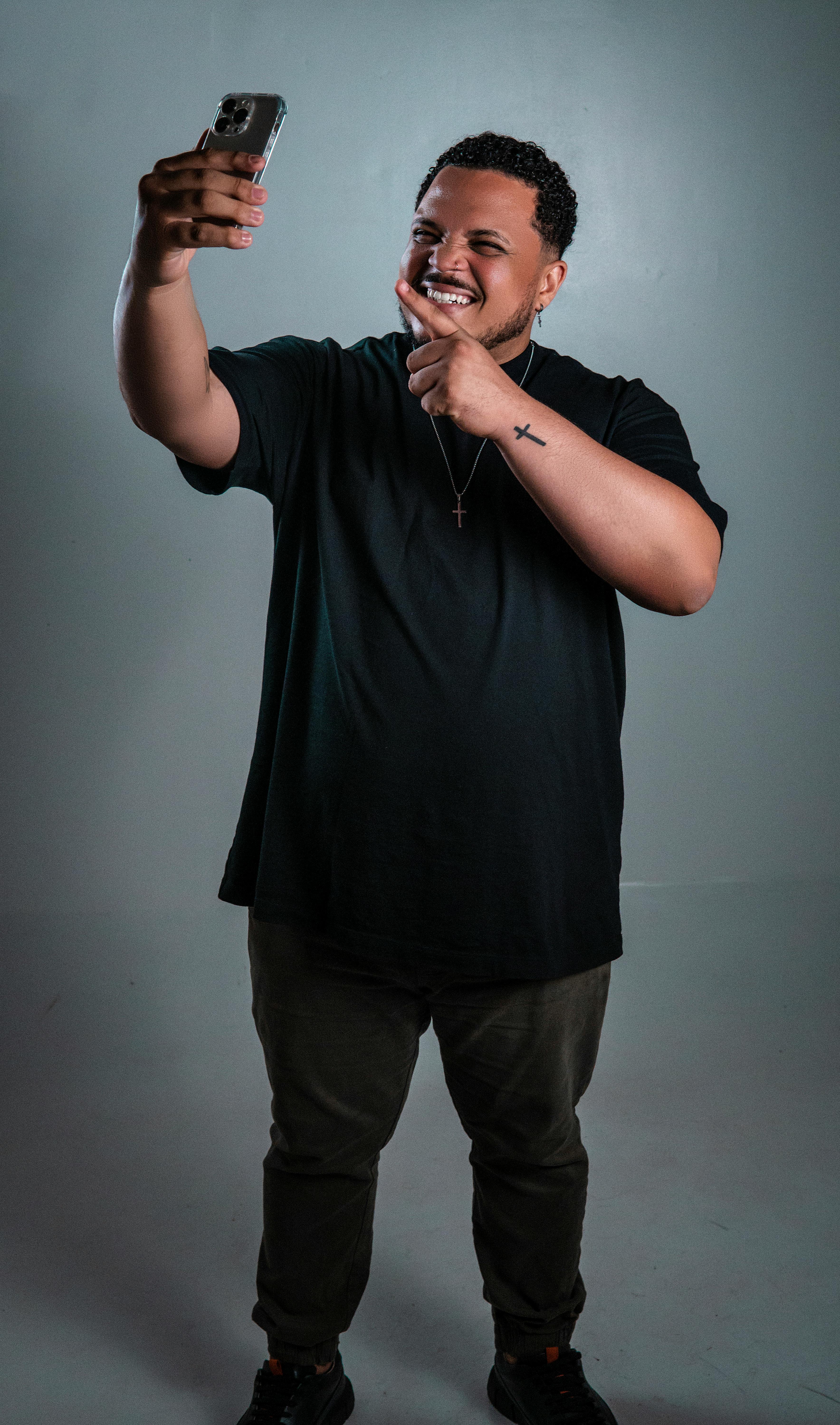 Smiling man in black t-shirt taking a selfie in a studio with gray background.