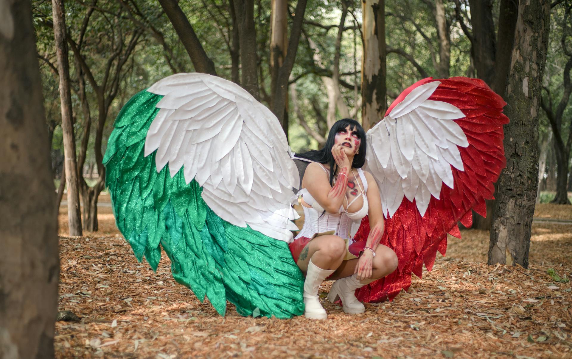 A woman in an elaborate angel costume squatting in a forest.