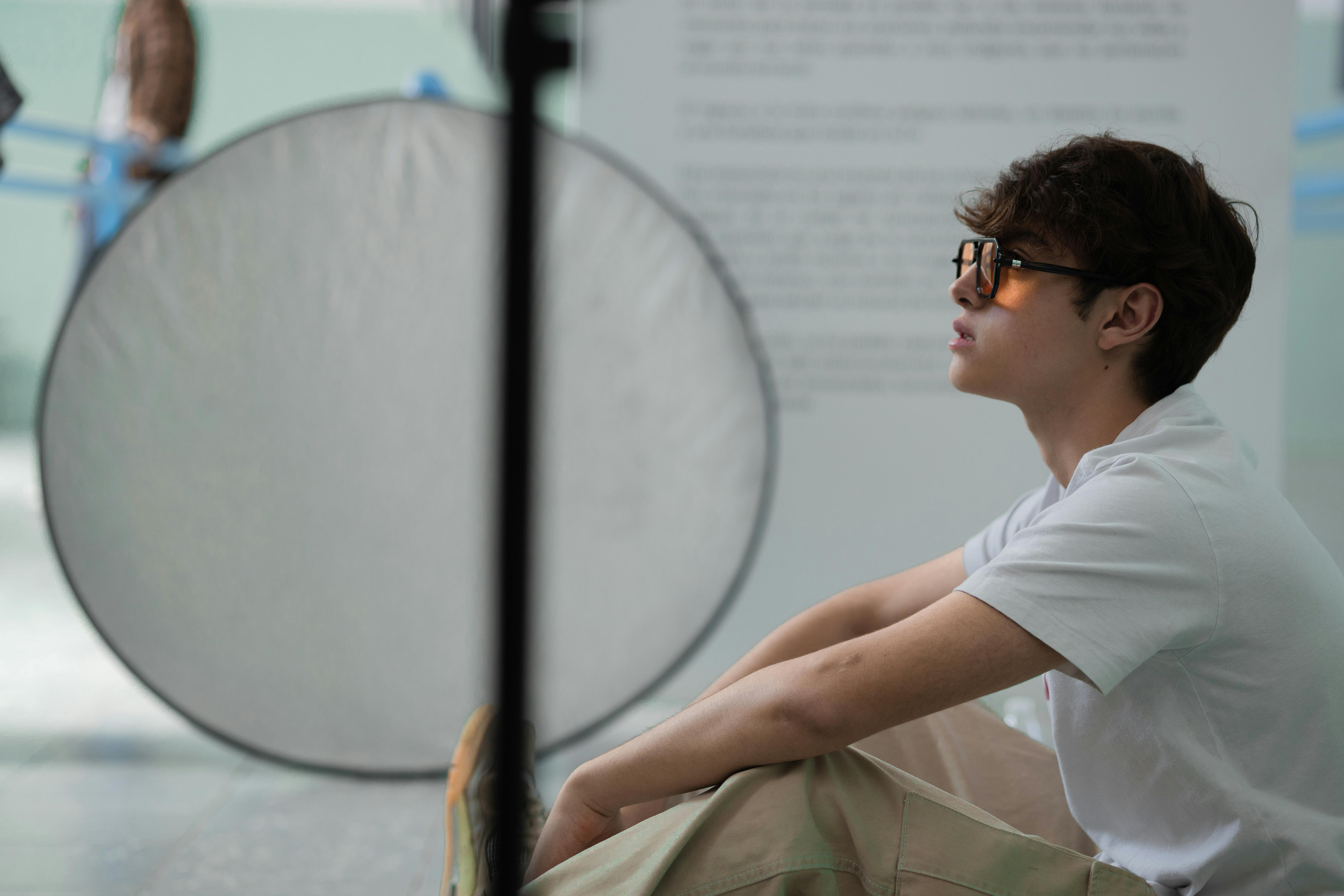 Stylish man wearing eyeglasses and white t-shirt sitting indoors in a relaxed pose.
