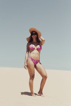 A woman poses in a bikini and sunhat on a sunny beach day, exuding summer vibes.