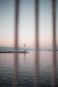 黄昏时分，灯塔在平静海水的环绕中显得格外宁静，天空泛着粉红色，前景有些模糊