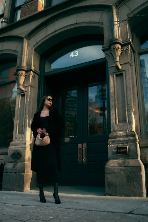 A woman in a black dress and hat stands outside a building