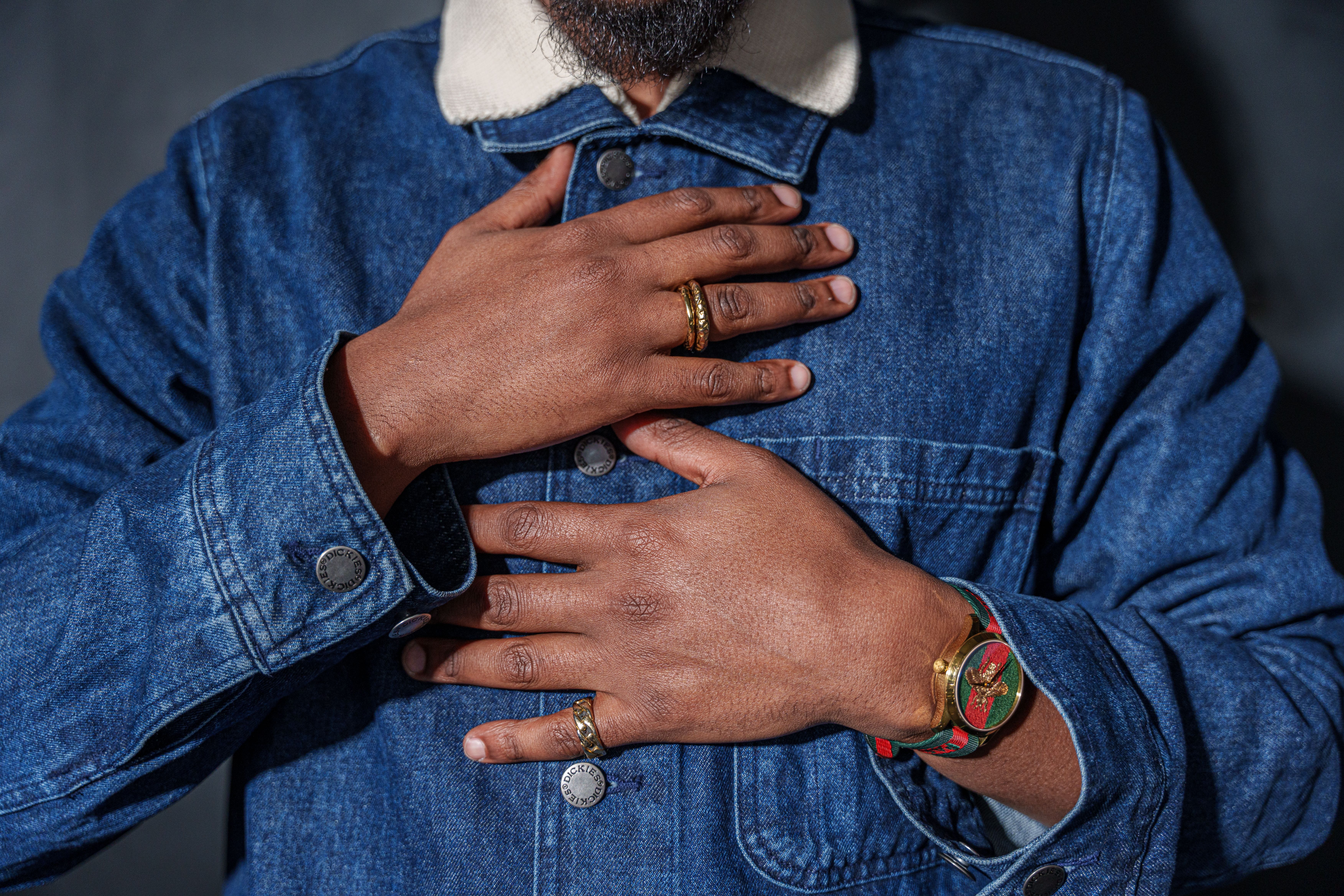 Close-up of a man in denim holding his hands over his chest, showcasing rings and a stylish watch.