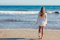 一名年轻女子身穿白色连衣裙，沿着阳光明媚的海滩漫步，欣赏着海景