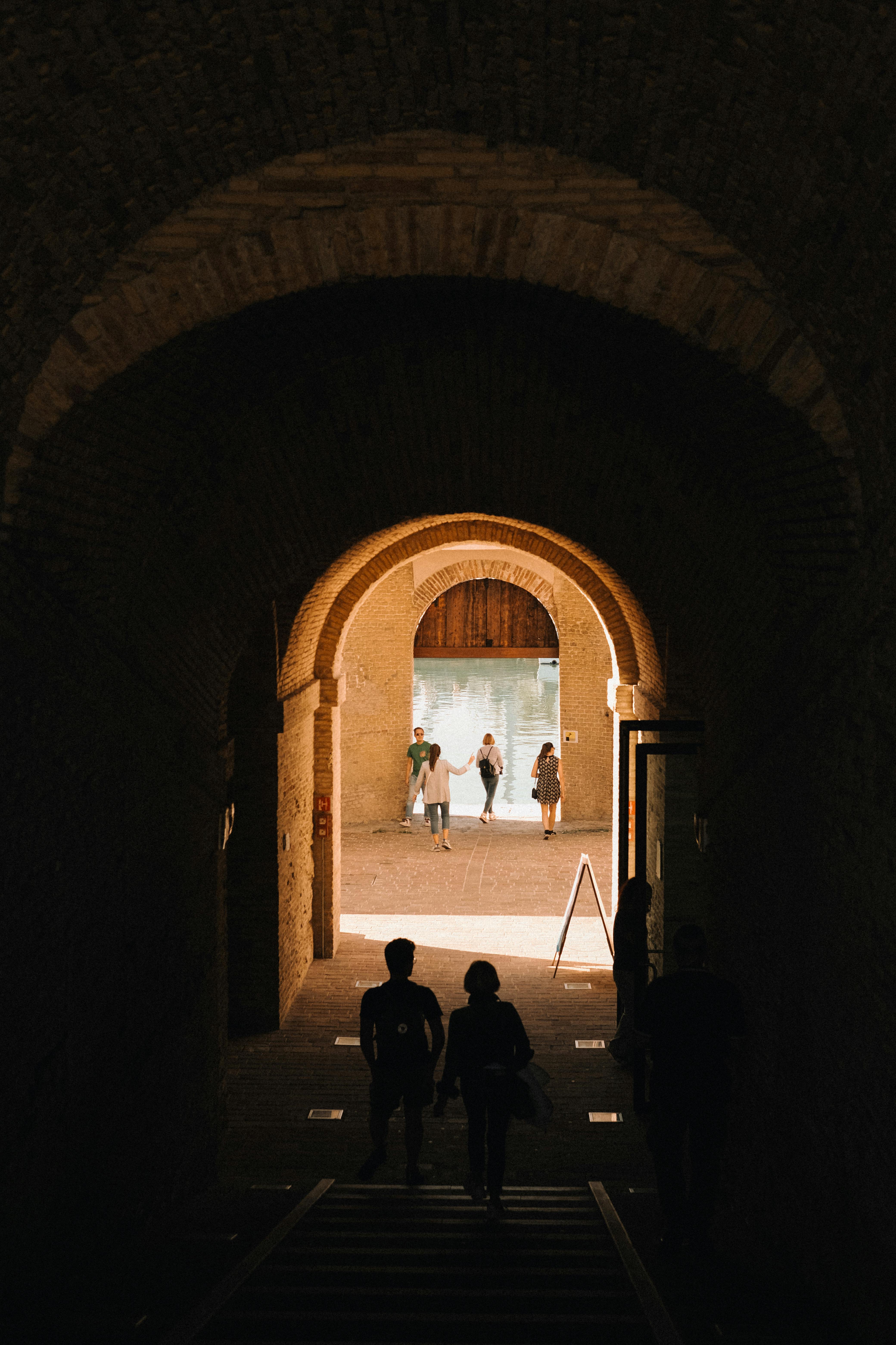 Silhouettes of people walking through a historic stone tunnel, backlit by warm sunlight.