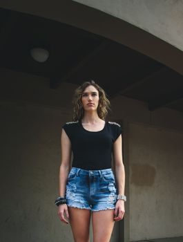 Portrait of a stylish young woman in denim shorts and black top standing under an architectural archway.