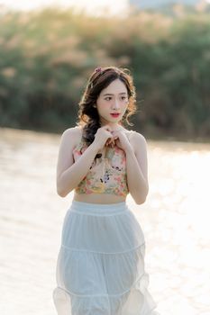 A young woman stands gracefully by the shimmering lake under the summer sun, showcasing fashion and beauty.