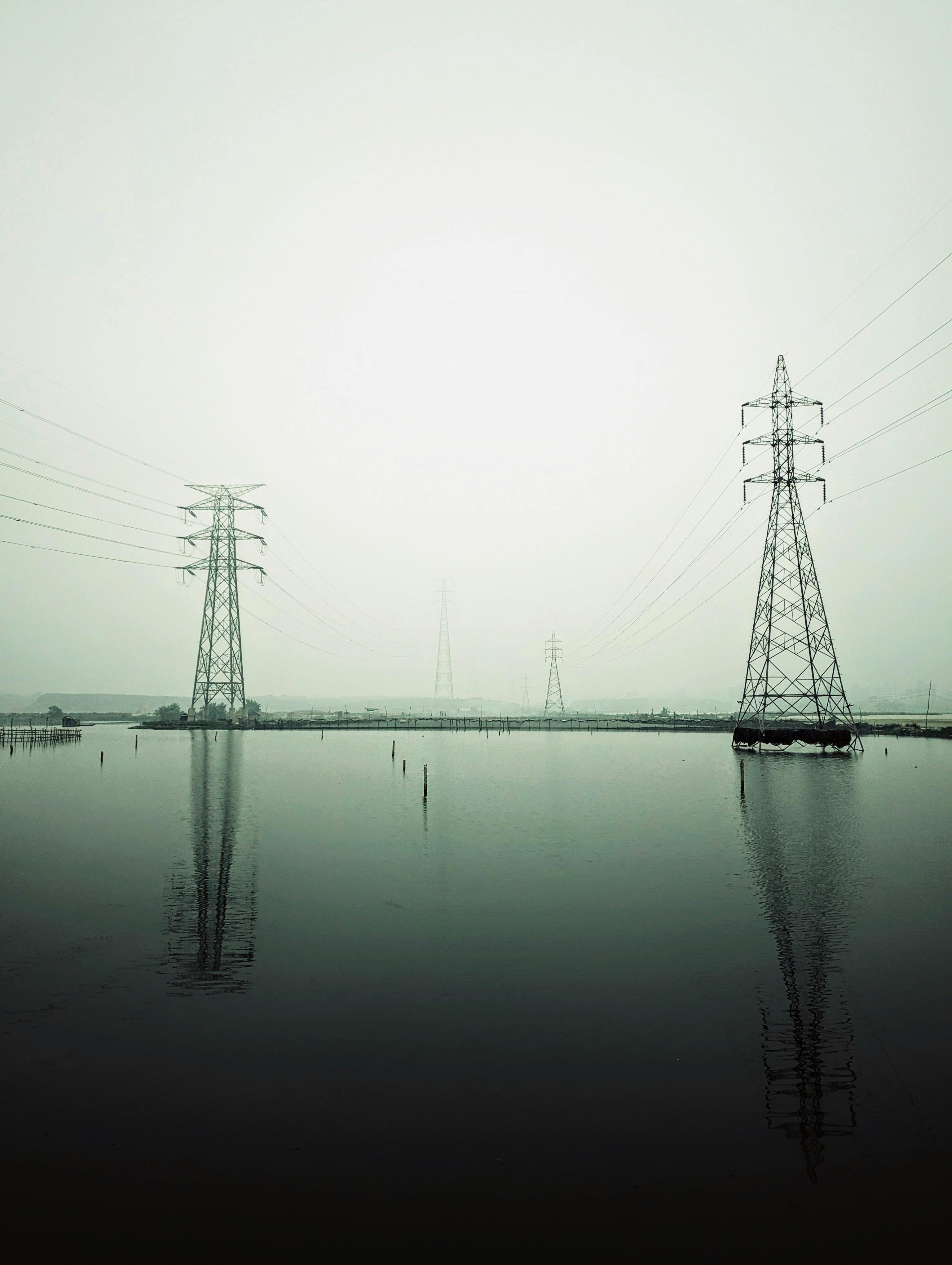 A tranquil scene of power lines towering over calm water reflecting a soft sunset glow.