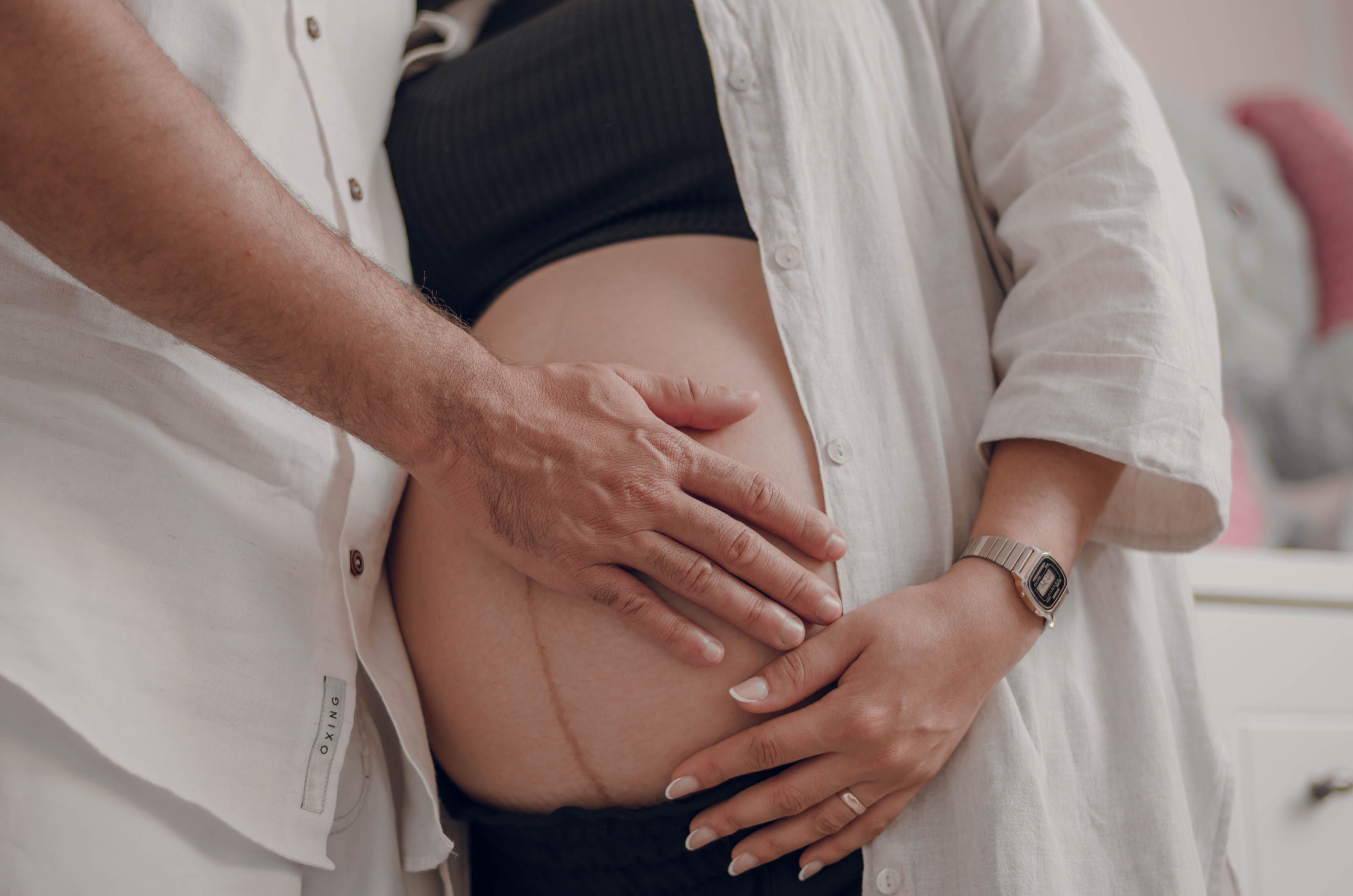 Close-up of a pregnant couple in white shirts lovingly touching a baby bump indoors.