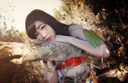 Elegant young woman leaning on a tree trunk, surrounded by vibrant foliage.