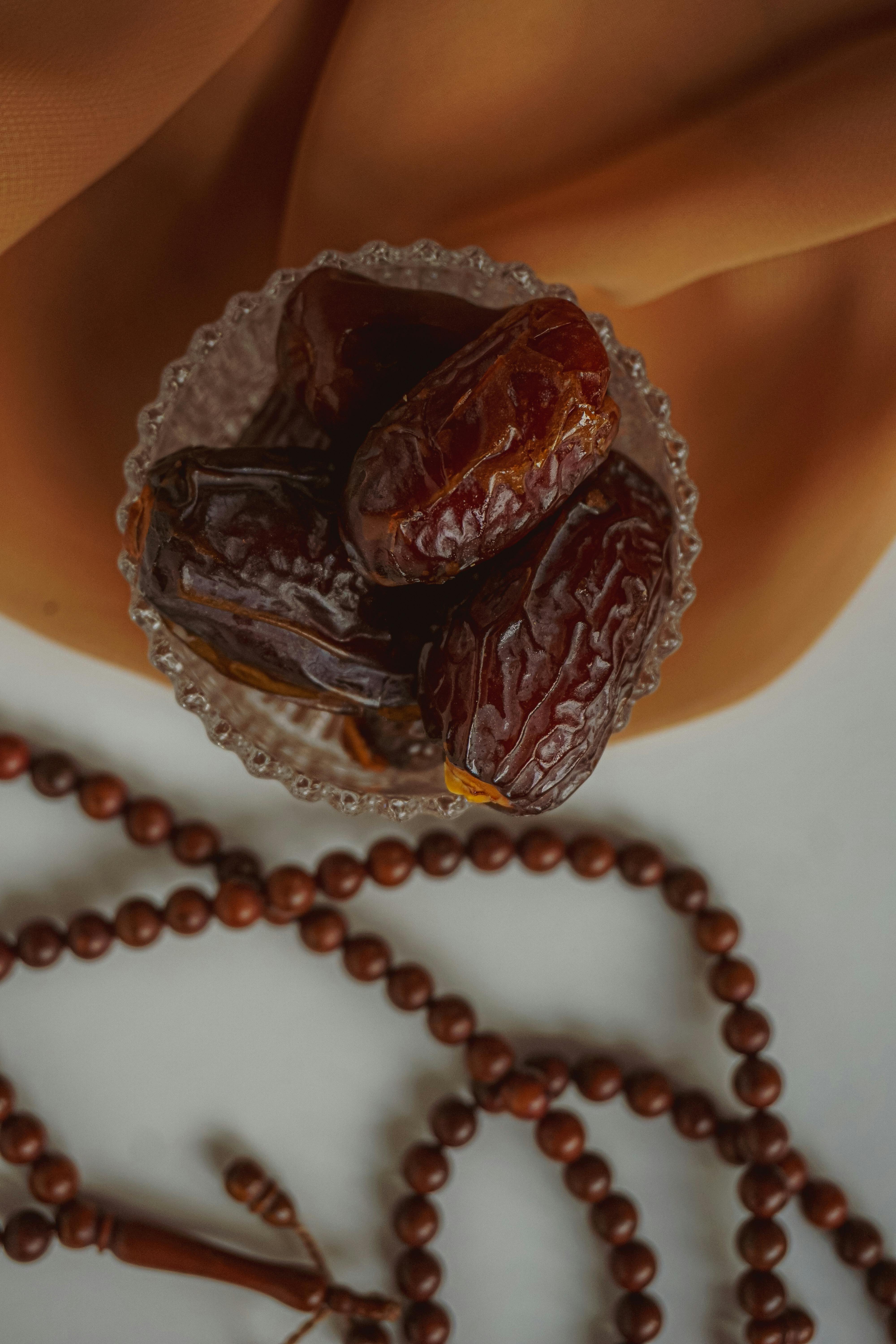 Close-up shot of dried dates in a clear bowl with beaded necklace on a fabric backdrop.
