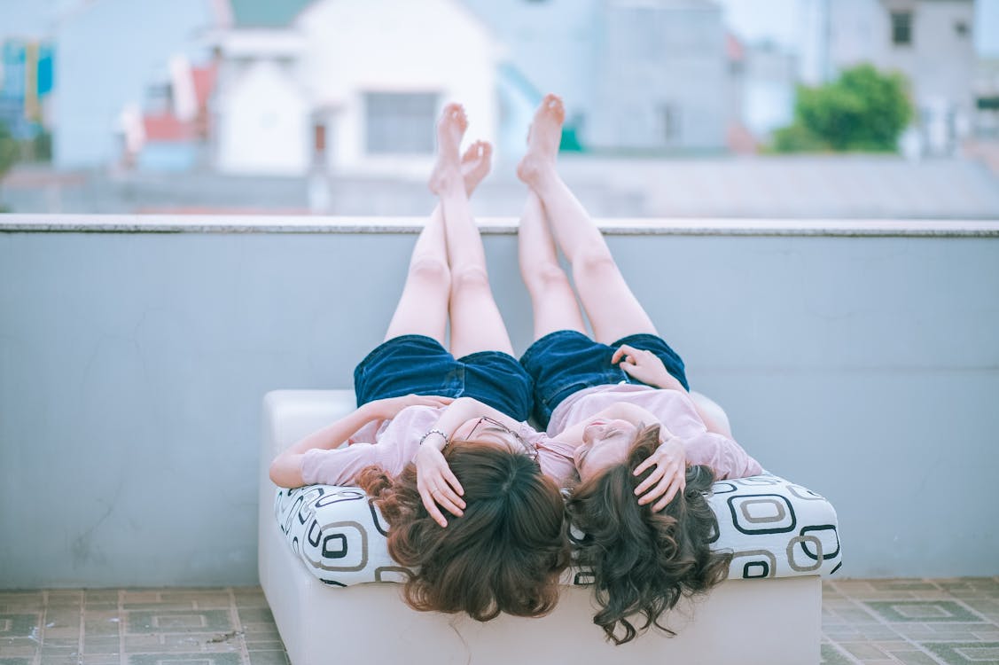 兩個女人穿著粉紅色的襯衫和藍色的短褲，躺在沙發上彼此面對，而雙腳交叉並放置在混凝土牆上