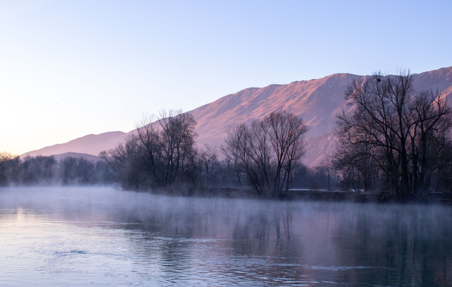 Still river at dusk below a pastel mountain ridge, mist rising off the water — photo by Mihailo Jovicevic on Pexels