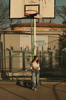 A young woman in jeans and crop top stands under a basketball hoop in an urban setting in Ankara, Türkiye.