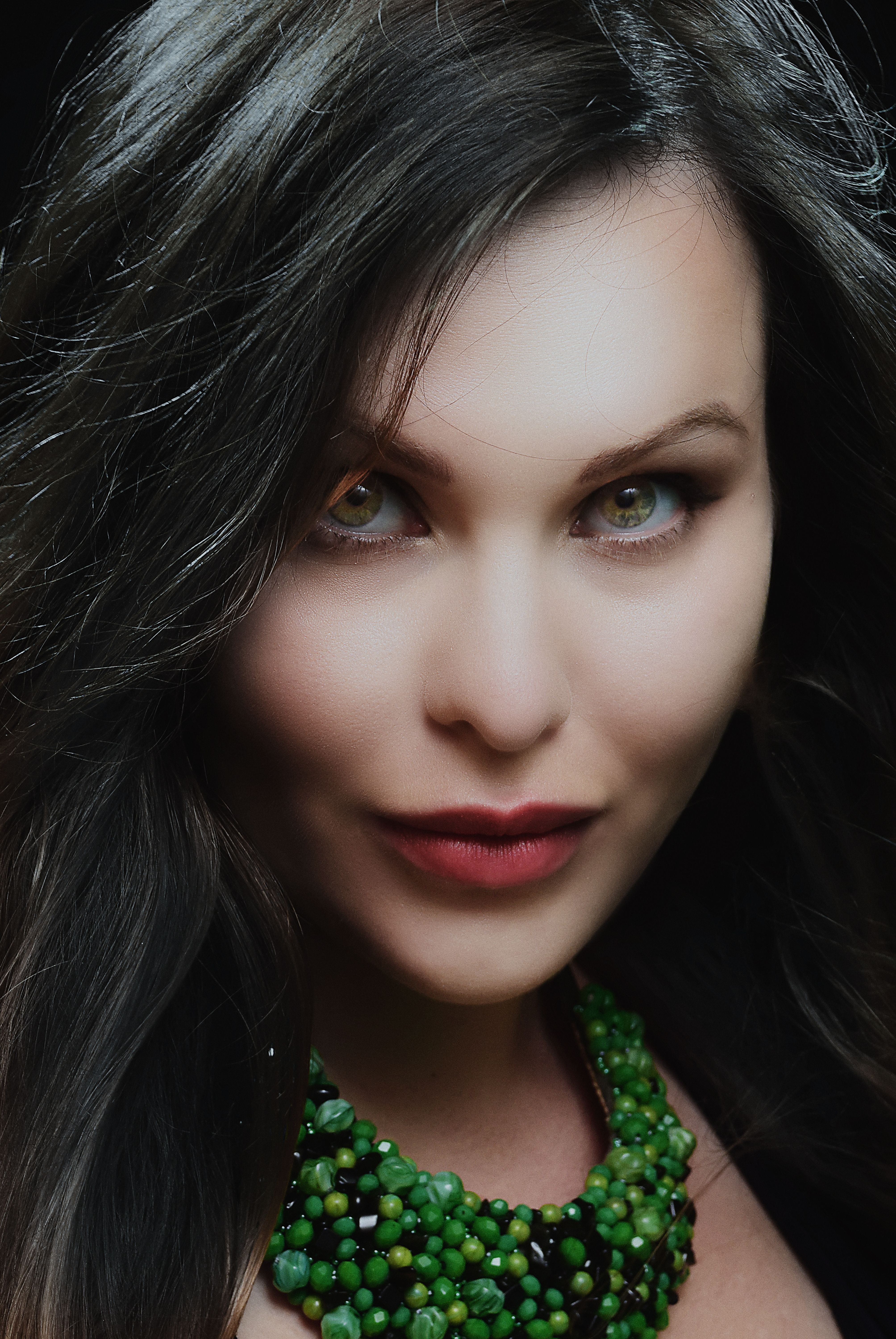 Close-up portrait of a brunette woman with striking green eyes and a green necklace in a studio setting.