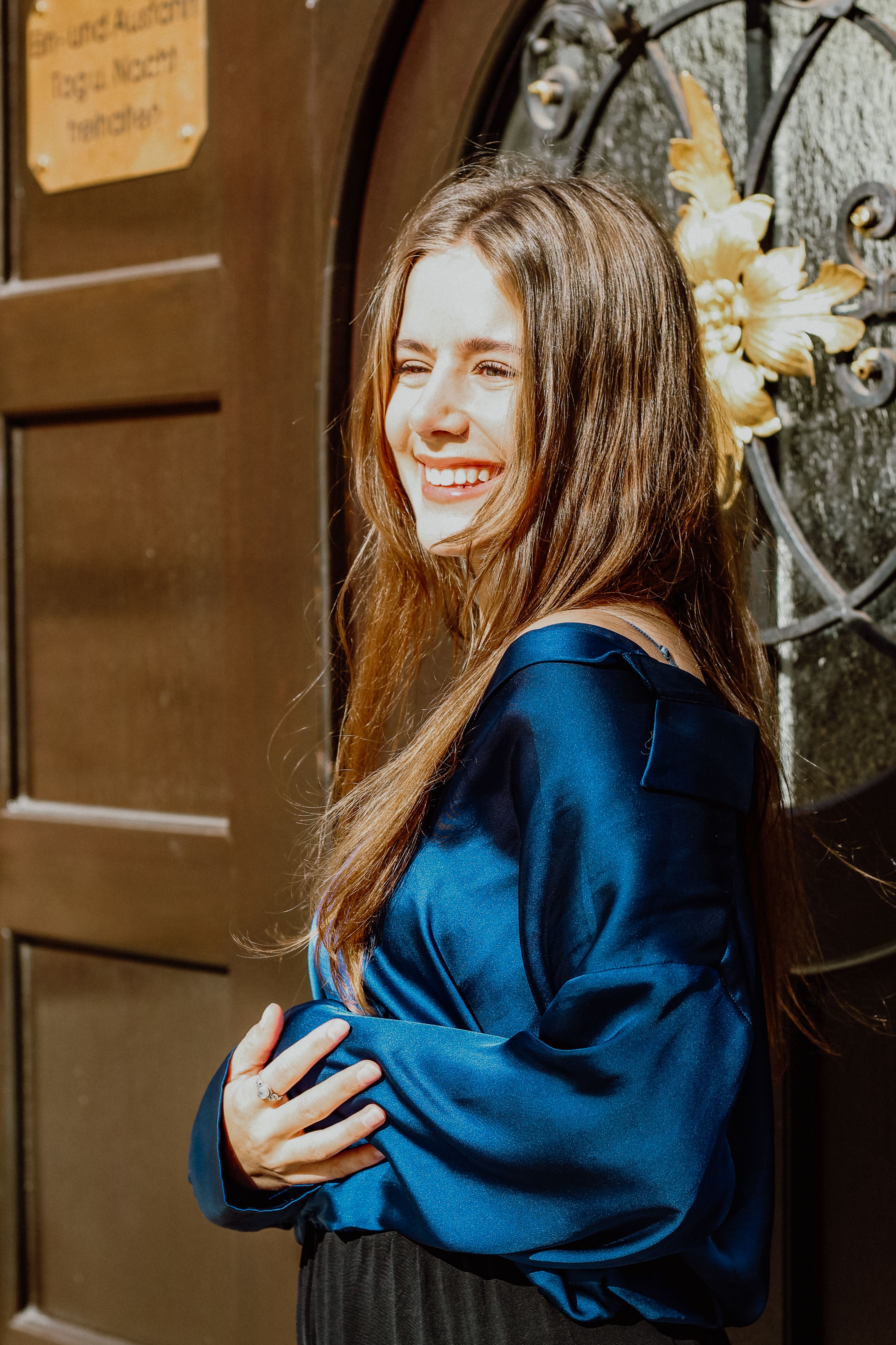 A joyful brunette woman smiling in a stylish blue satin blouse standing outdoors in sunlight.