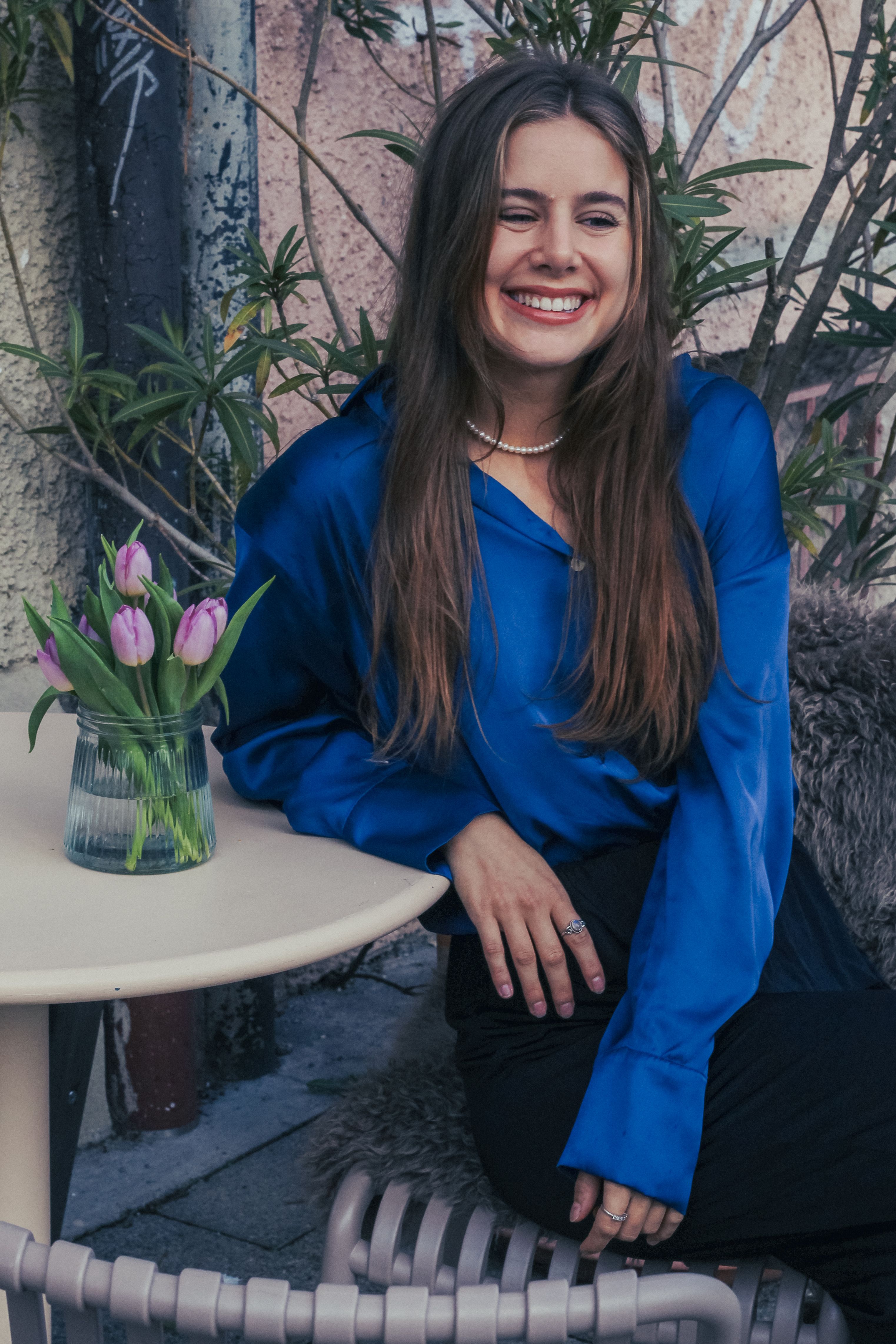 A cheerful woman sitting outside in a blue shirt, with purple tulips on the table.