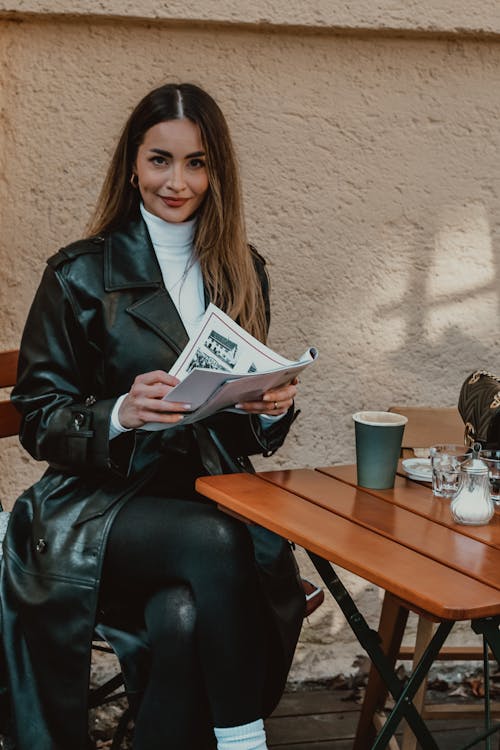 A woman in black leather jacket sitting at a table reading a magazine