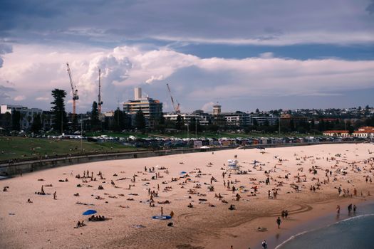 A bustling beach with people relaxing and a city skyline with construction cranes.
