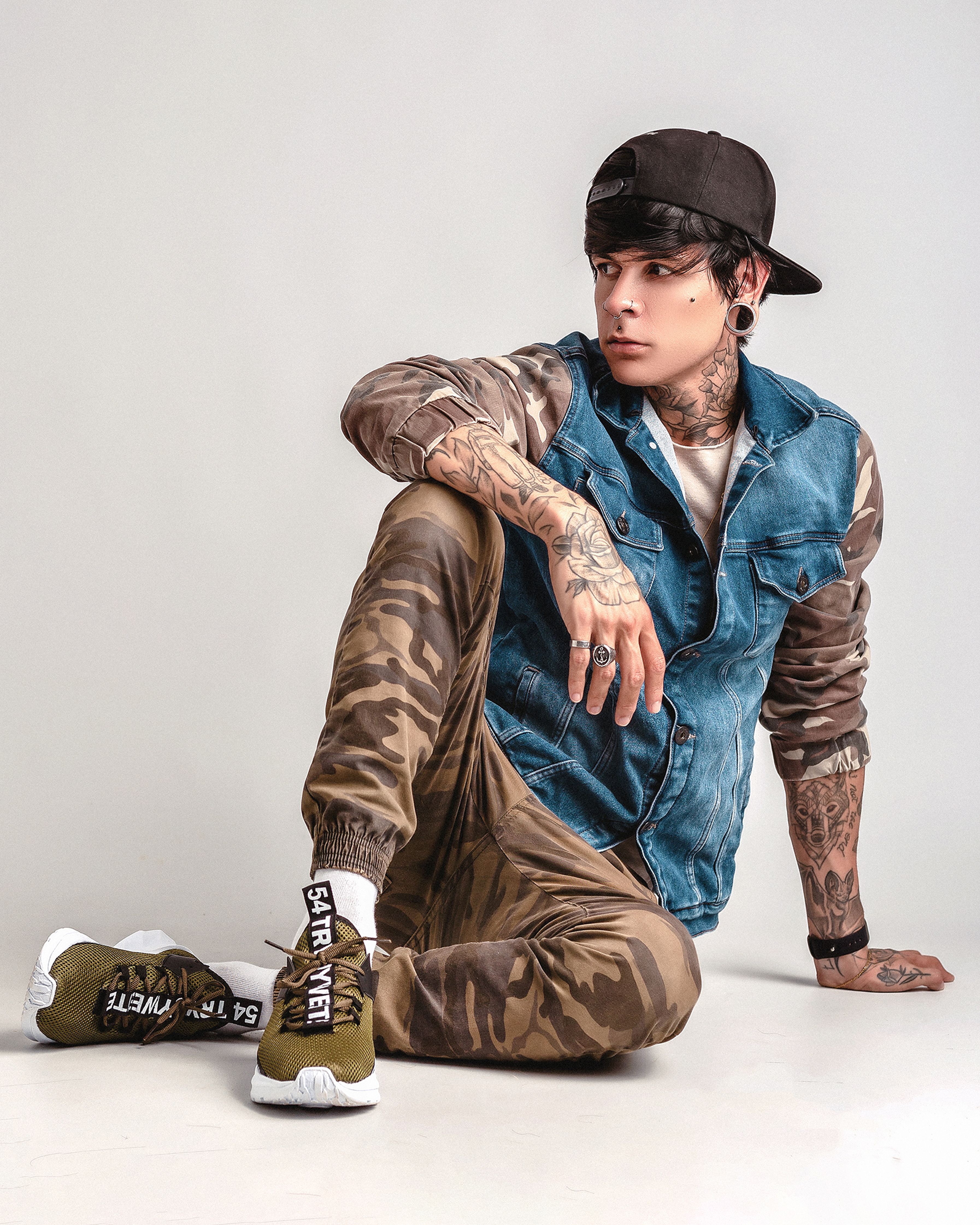 Stylish tattooed man in denim vest and camo pants sitting on white backdrop, showcasing urban fashion.