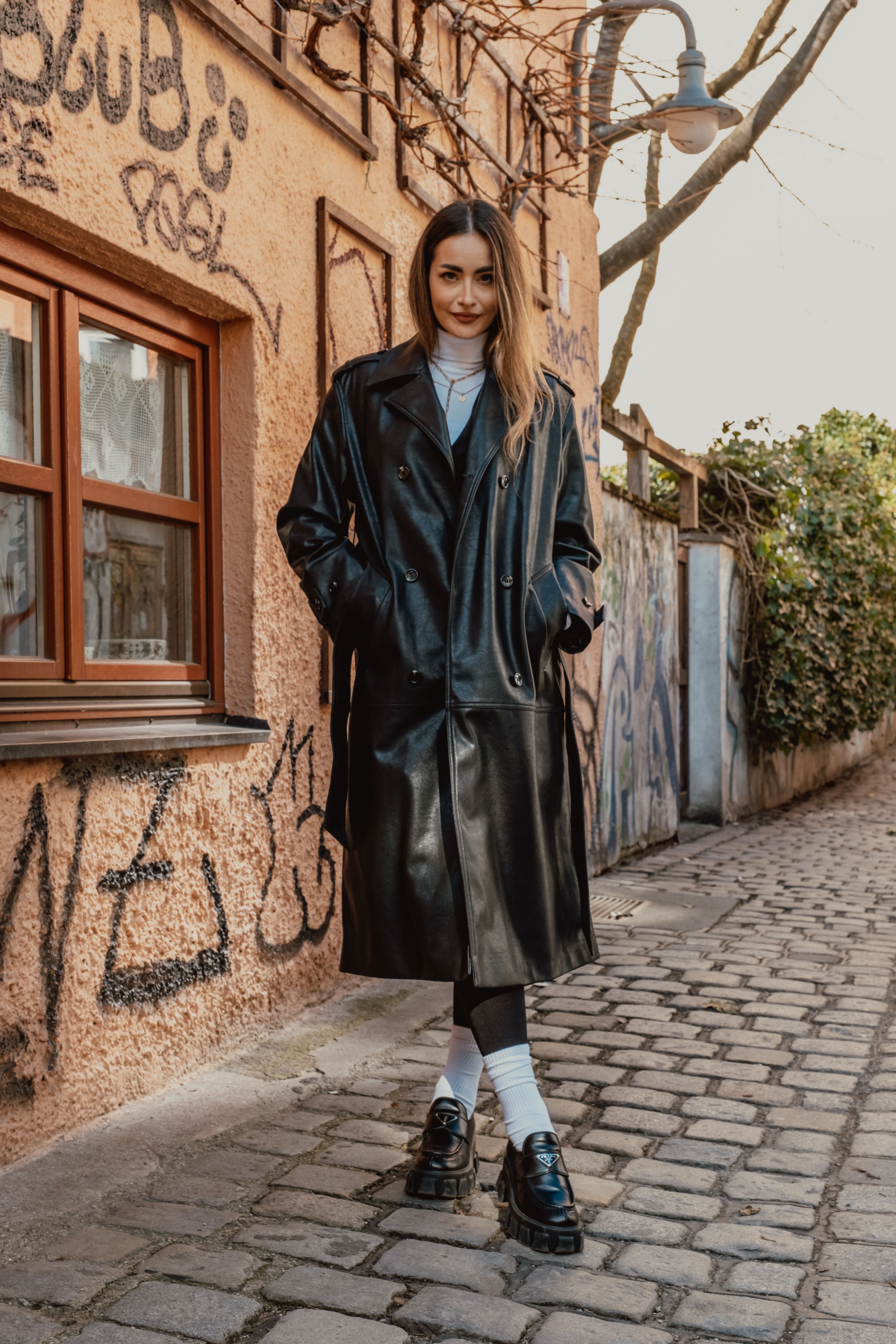 Stylish woman posing in a leather coat on a graffiti-lined street, showcasing urban fashion.