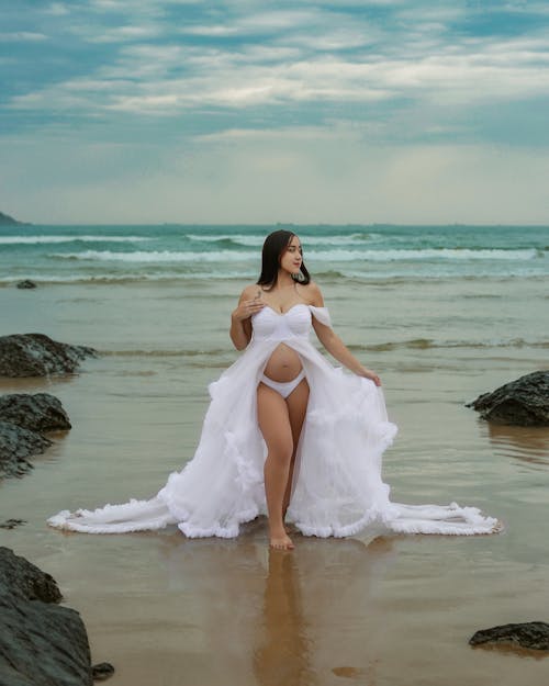 A pregnant woman in a white dress standing on the beach
