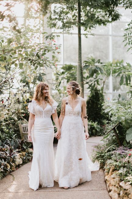 Two brides holding hands in a lush garden, showcasing elegance and joy.