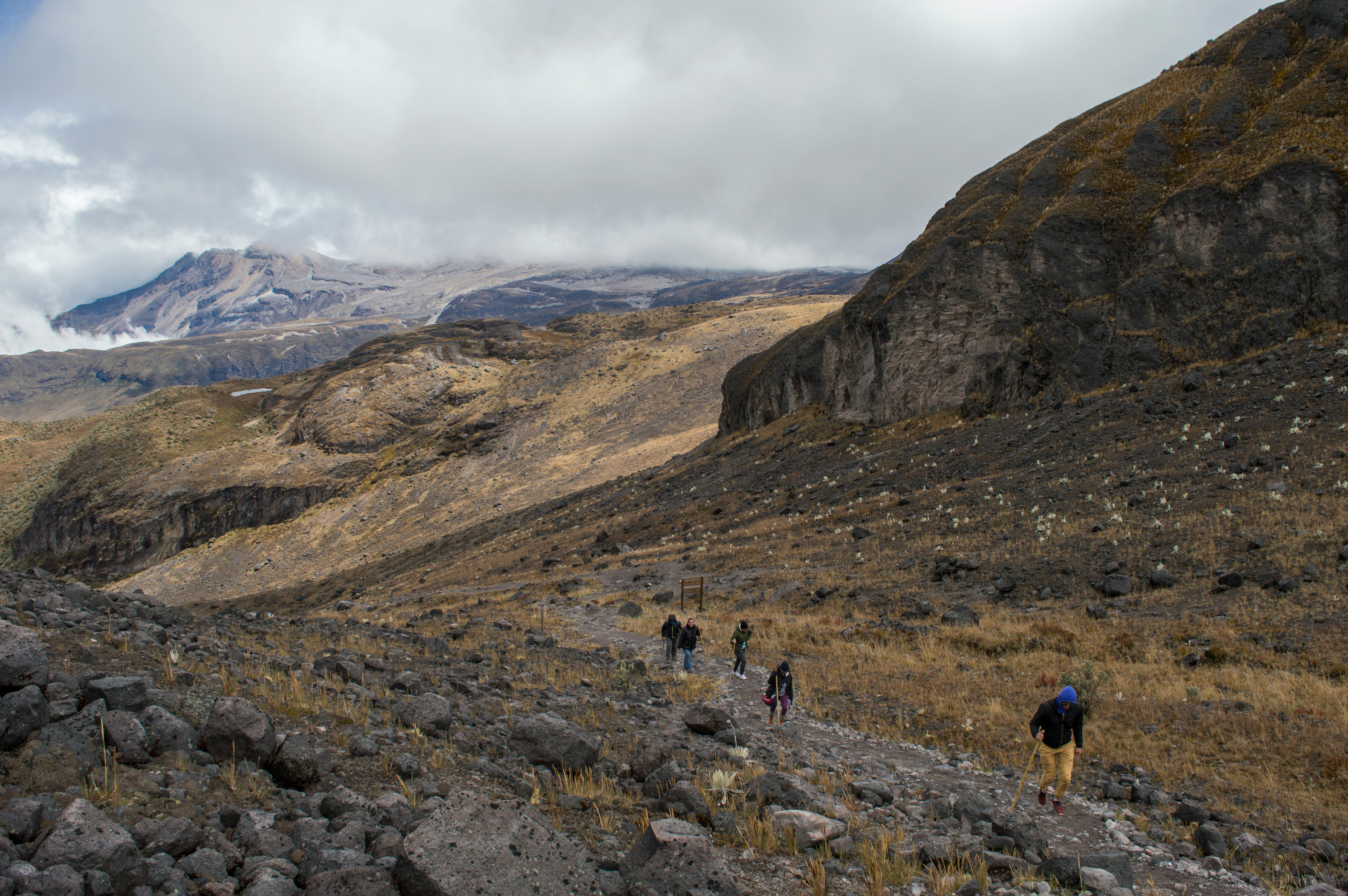 A group of hikers traverses a rugged mountain path under cloudy skies.