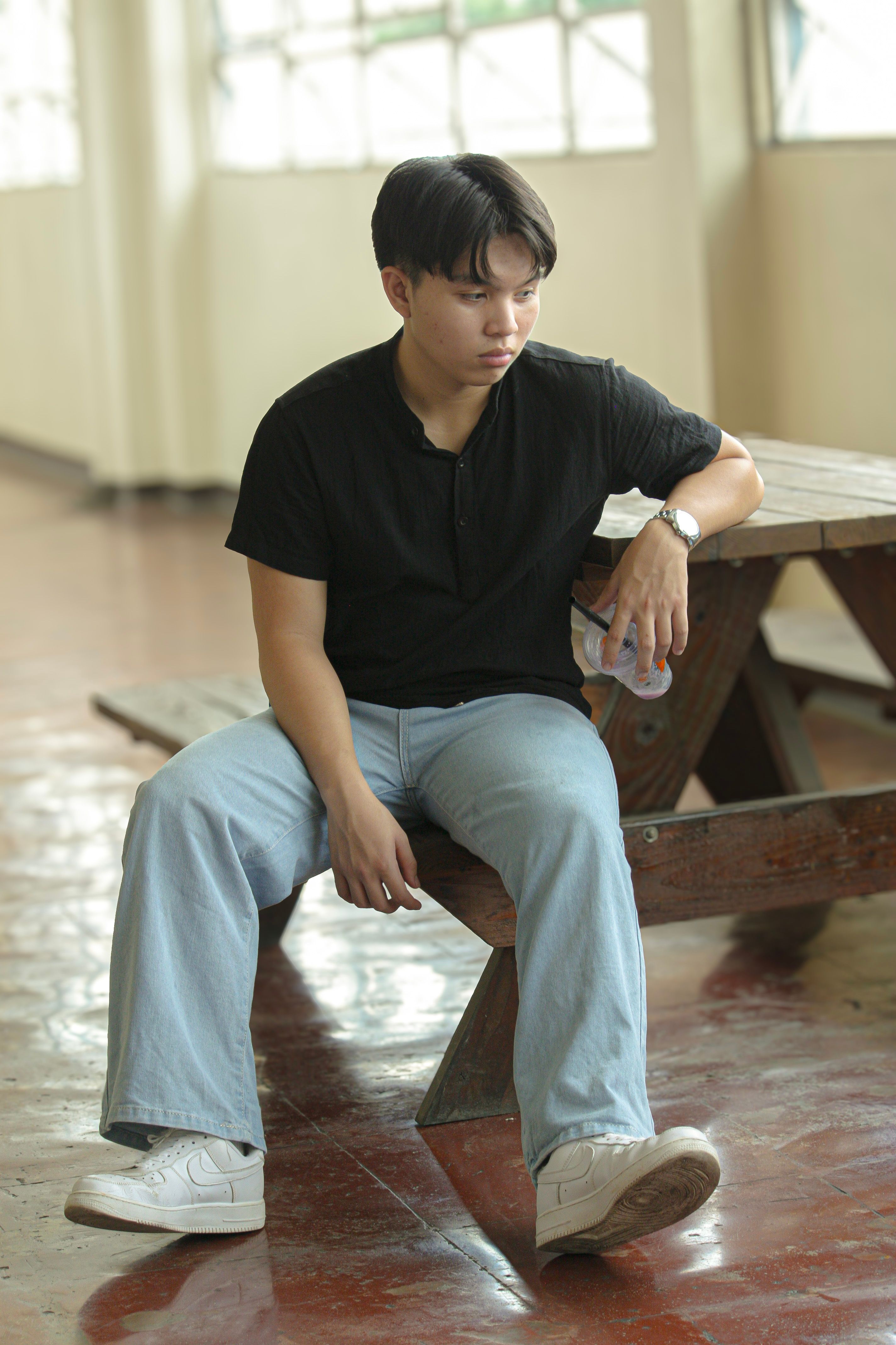 Portrait of a young man seated indoors on a wooden bench, dressed casually in a t-shirt and jeans.