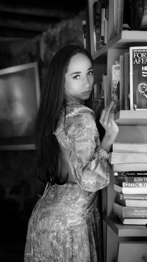 A woman in a dress leaning against a bookcase