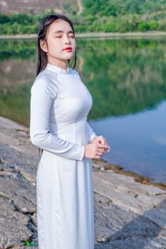 Asian woman in a white traditional dress with eyes closed, enjoying the serene outdoor setting by the water.