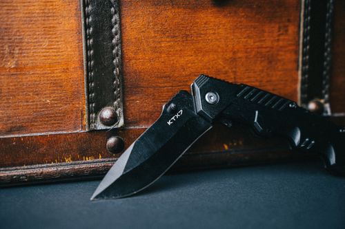 Free Detailed close-up of a black folding knife with a wooden background, highlighting its sharp blade and sturdy design. Stock Photo