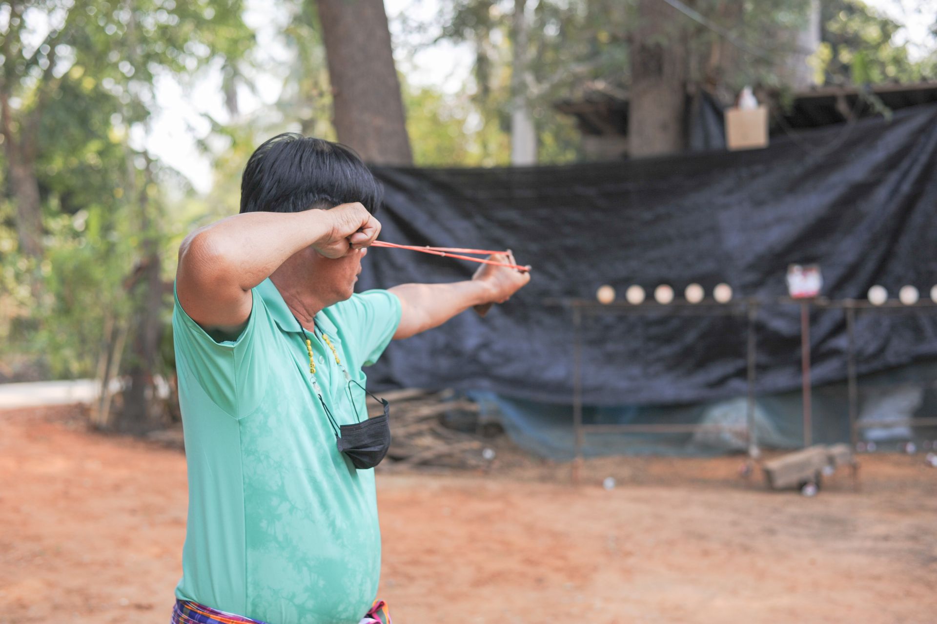 An adult man aiming with a slingshot at targets in an outdoor park setting.