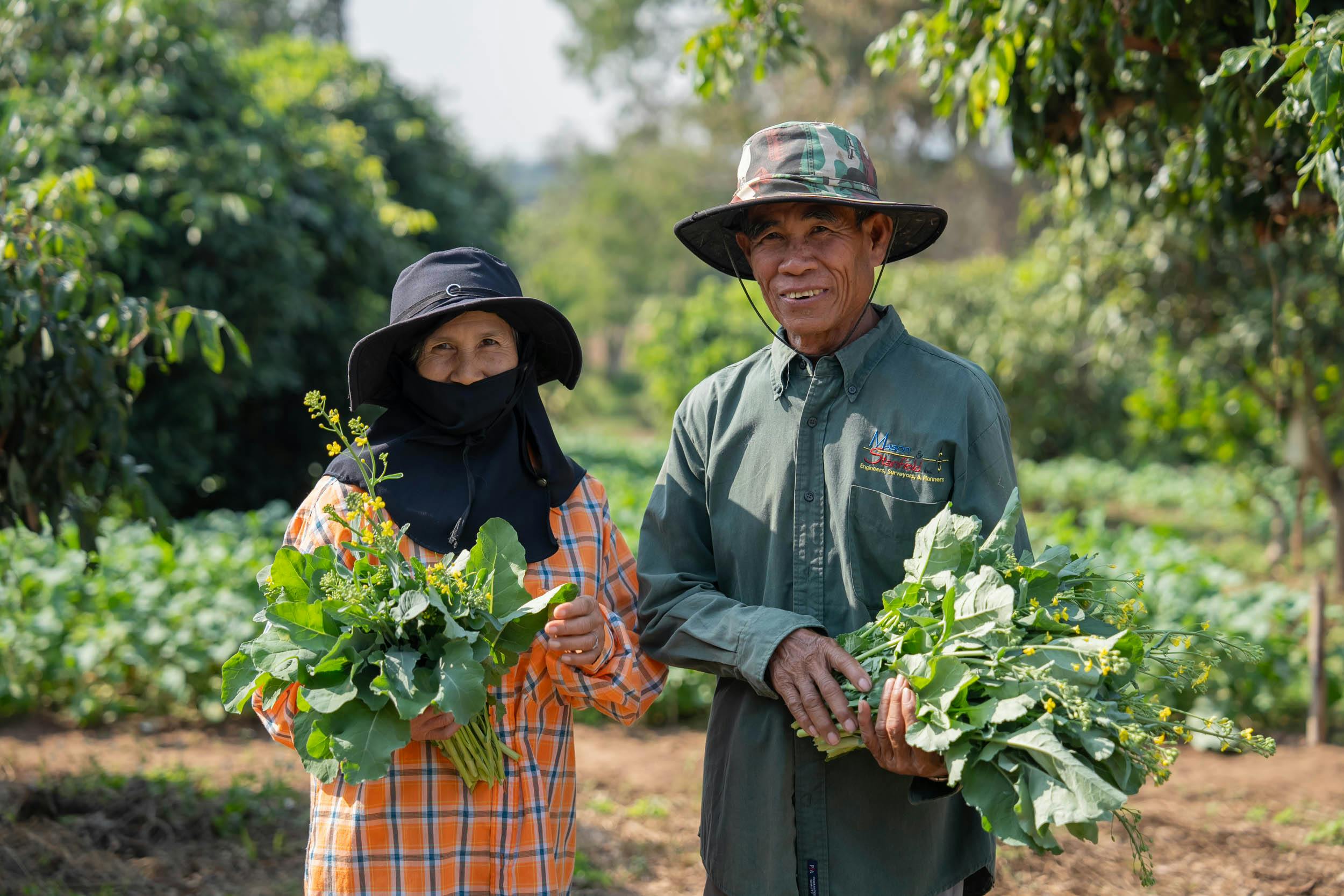 Elderly Asian couple smiling and holding freshly harvested greens in a rural farm setting.