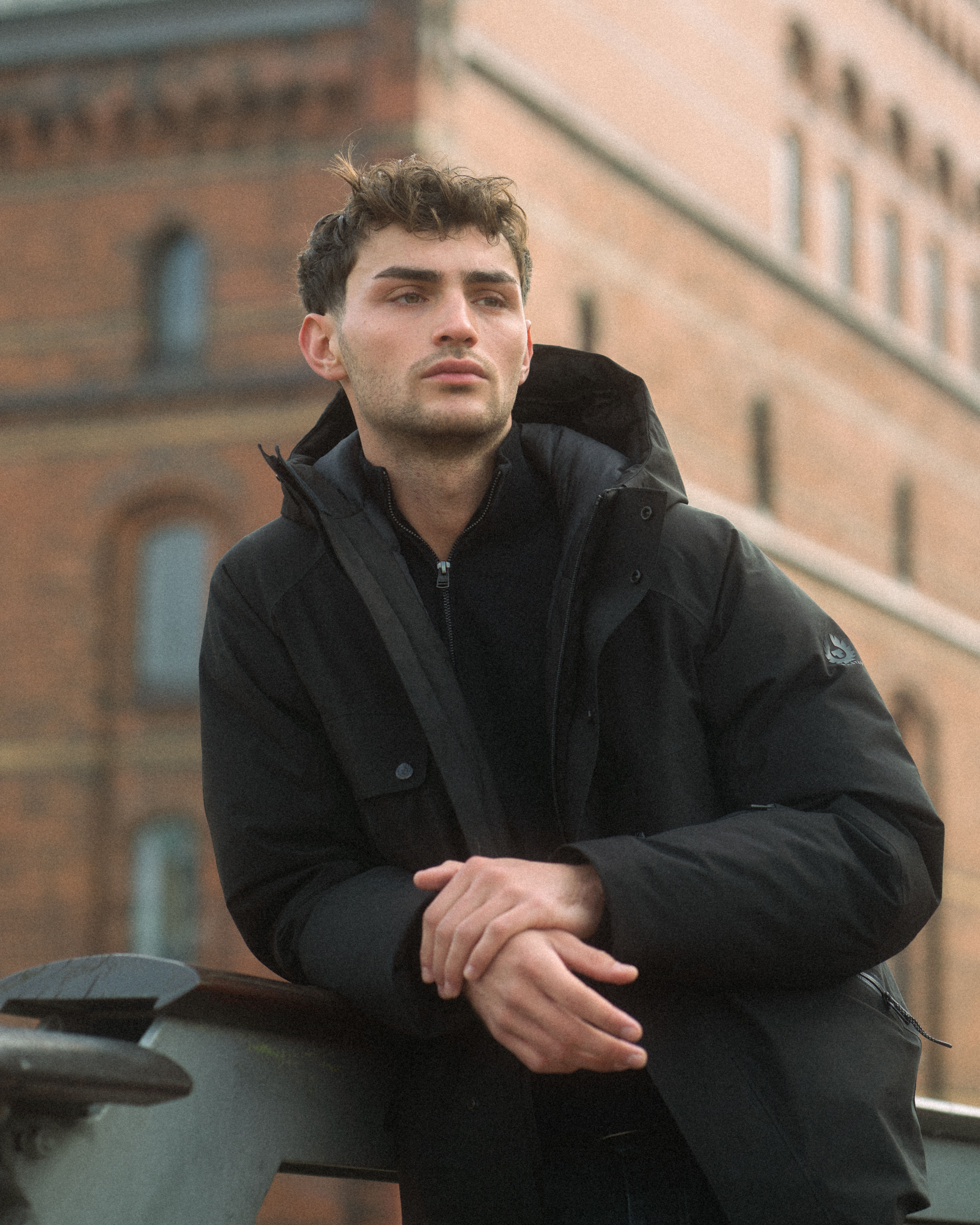 A fashionable man posing in a black jacket against a brick building backdrop.