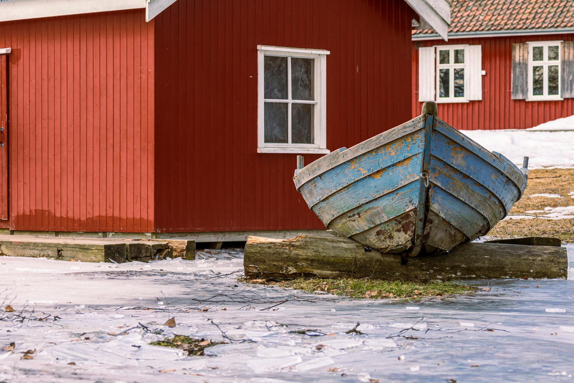 A serene winter scene of a rustic red house with a vintage blue boat on icy ground.